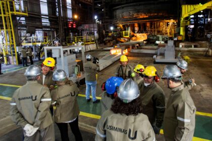 Trabalhadores usam capacetes e uniformes de segurança em ambiente industrial da Usiminas, com máquinas e estruturas metálicas ao fundo.