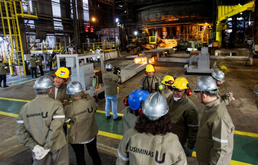 Trabalhadores usam capacetes e uniformes de segurança em ambiente industrial da Usiminas, com máquinas e estruturas metálicas ao fundo.