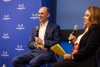 Dois palestrantes sorridentes em um palco azul, segurando microfones e notas, discutem temas importantes em evento.