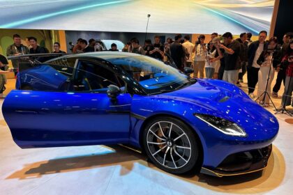 Carro esportivo azul em exibição, com porta aberta, cercado por pessoas tirando fotos em um salão de automóveis.