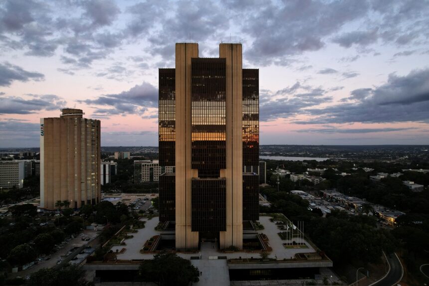 Prédio Central do Banco Central em Brasília ao entardecer, refletindo o céu colorido, com edifícios e árvores ao redor.