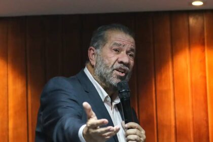 Orador de terno fala ao microfone em uma sala madeira, gesticulando, transmitindo mensagem com expressão enfática.
