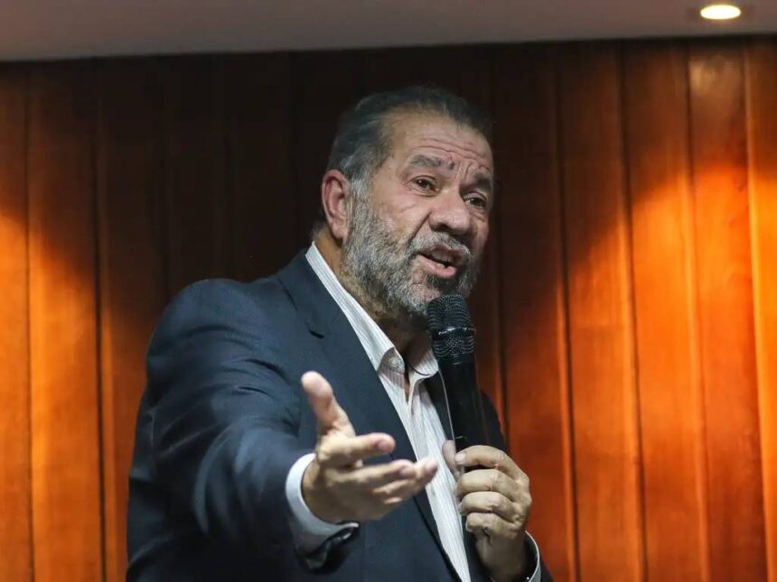 Orador de terno fala ao microfone em uma sala madeira, gesticulando, transmitindo mensagem com expressão enfática.