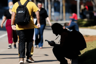Pessoa agachada usando celular em calçada movimentada, com pedestres ao fundo. Cena urbana em dia ensolarado.