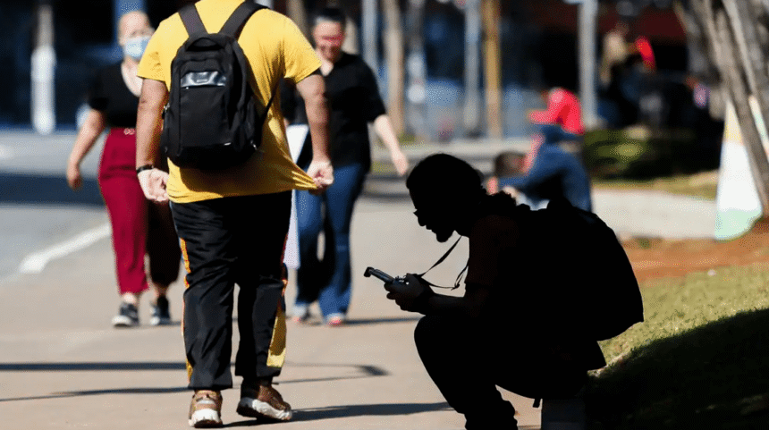 Pessoa agachada usando celular em calçada movimentada, com pedestres ao fundo. Cena urbana em dia ensolarado.