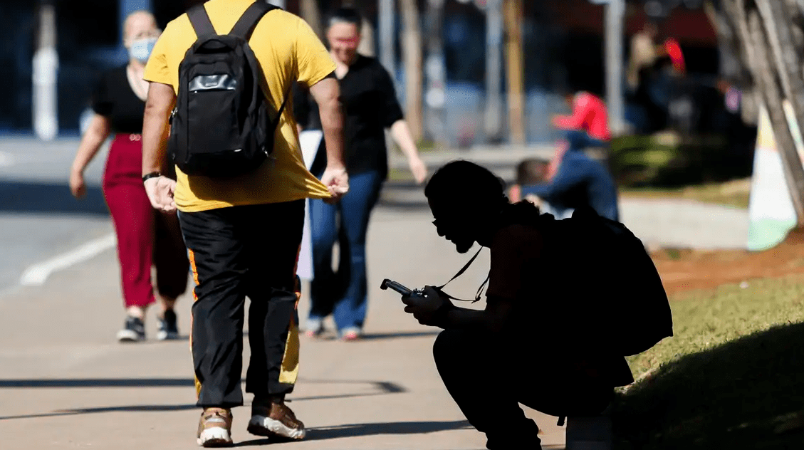 Pessoa agachada usando celular em calçada movimentada, com pedestres ao fundo. Cena urbana em dia ensolarado.