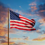 Bandeira dos Estados Unidos ondulando ao vento, destacando-se contra um pôr do sol vibrante com nuvens coloridas.