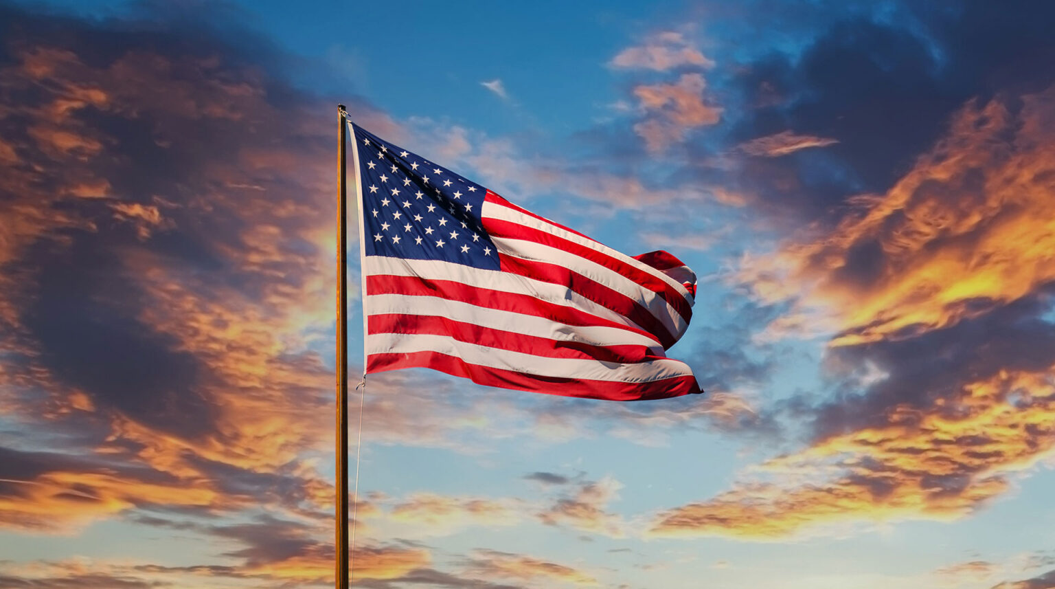 Bandeira dos Estados Unidos ondulando ao vento, destacando-se contra um pôr do sol vibrante com nuvens coloridas.