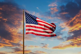 Bandeira dos Estados Unidos ondulando ao vento, destacando-se contra um pôr do sol vibrante com nuvens coloridas.