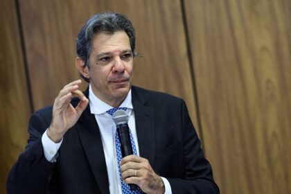 Homem em discurso, vestindo terno escuro e gravata azul, gesticula com a mão enquanto segura microfone em palco de madeira.