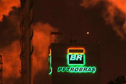 Refinaria da Petrobras à noite, com logotipo iluminado em destaque e fumaça densa emitida, criando um cenário industrial.