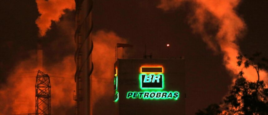 Refinaria da Petrobras à noite, com logotipo iluminado em destaque e fumaça densa emitida, criando um cenário industrial.