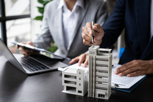 Duas pessoas em reunião analisam contratos imobiliários, com laptop e maquetes de prédios sobre a mesa.