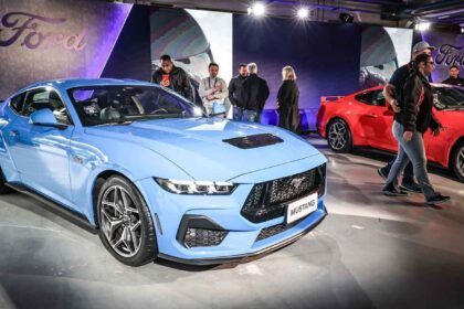 Pessoas observam novas edições do Ford Mustang em um evento, destacando modelos em azul e vermelho sob iluminação especial.