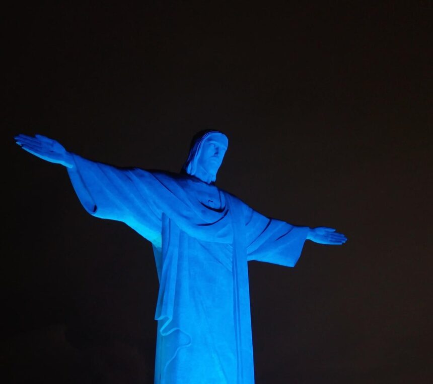 Estátua do Cristo Redentor iluminada de azul à noite, destacando-se contra o céu escuro no Rio de Janeiro.