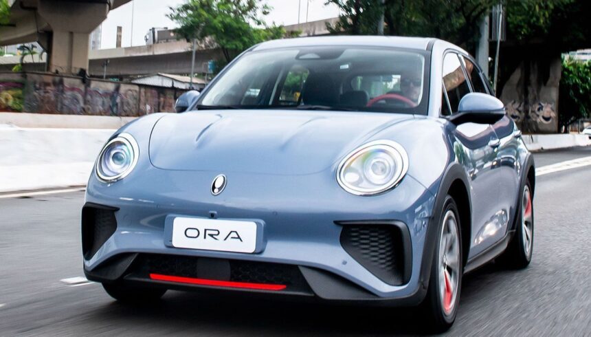 Carro elétrico ORA cinza circula em rua urbana, destacando design moderno e eficiente para mobilidade sustentável.