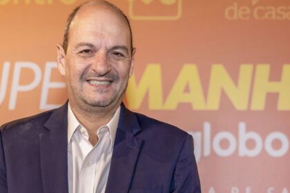 Homem sorridente de terno azul e camisa branca, em frente a um fundo laranja com textos promocionais desfocados.