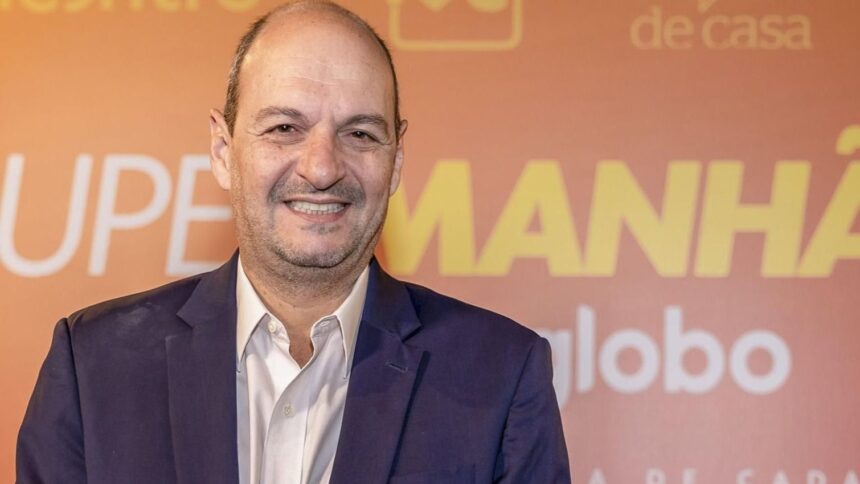 Homem sorridente de terno azul e camisa branca, em frente a um fundo laranja com textos promocionais desfocados.