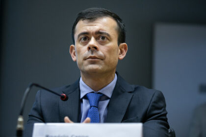 Homem de terno e gravata azul fala em microfone, participando de um evento ou conferência, com expressão séria.