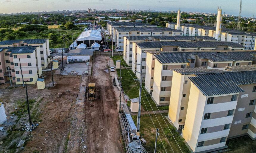 Complexo habitacional em construção com prédios residenciais ao lado de estrada de terra e maquinário pesado.