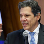 Homem de terno discursando, segurando microfone, com expressão séria e gesticulando enquanto fala em evento.