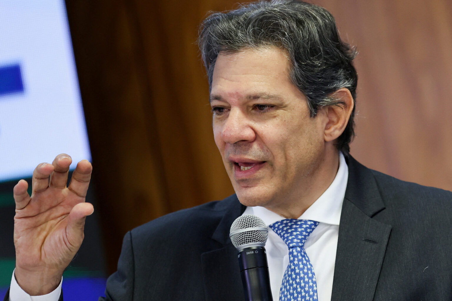 Homem de terno discursando, segurando microfone, com expressão séria e gesticulando enquanto fala em evento.