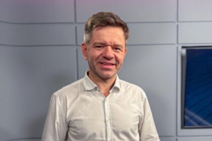 Homem com camisa branca sorri em um estúdio de televisão moderno com painéis cinza e azul ao fundo.