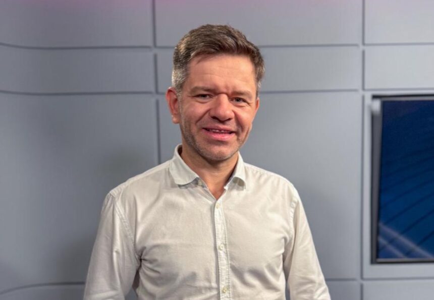 Homem com camisa branca sorri em um estúdio de televisão moderno com painéis cinza e azul ao fundo.