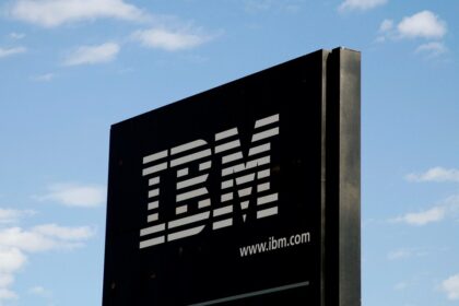 Placa da IBM com logo em fundo preto, destacando-se contra um céu azul com nuvens, enfatizando tecnologia e inovação.