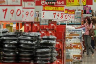 Clientes em loja de departamento, cercados por promoções e produtos domésticos, pesquisando preços e escolhendo itens.