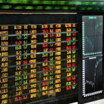 Painel eletrônico de cotações da bolsa de valores, exibindo números em cores variadas, gráficos de tendências ao lado.