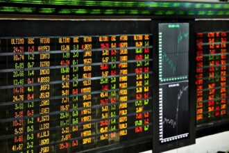 Painel eletrônico de cotações da bolsa de valores, exibindo números em cores variadas, gráficos de tendências ao lado.