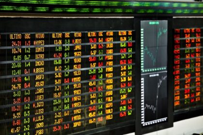 Painel eletrônico de cotações da bolsa de valores, exibindo números em cores variadas, gráficos de tendências ao lado.