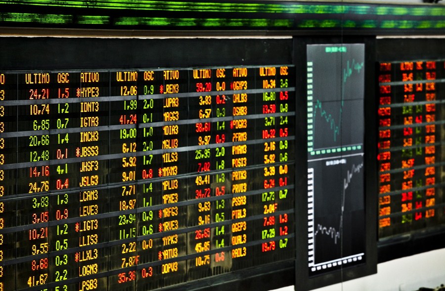 Painel eletrônico de cotações da bolsa de valores, exibindo números em cores variadas, gráficos de tendências ao lado.