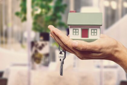 Mão segurando uma pequena casa em miniatura e uma chave, simbolizando compra ou aluguel de imóvel.