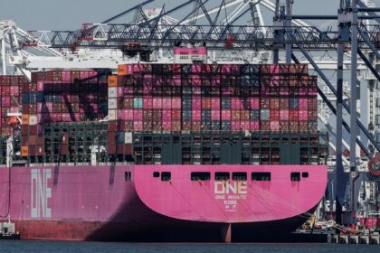 Navio cargueiro rosa com contêineres empilhados atracado em porto; guindastes ao fundo. Transporte marítimo global.