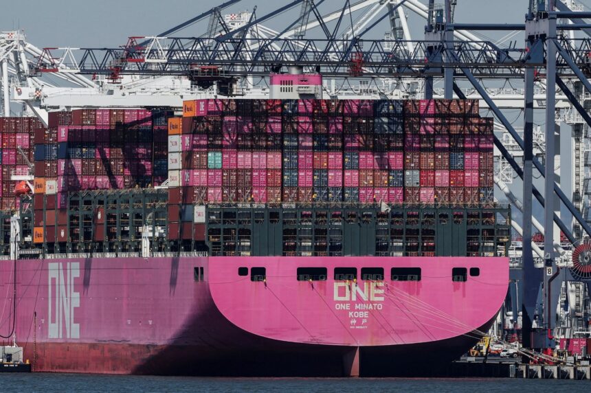 Navio cargueiro rosa com contêineres empilhados atracado em porto; guindastes ao fundo. Transporte marítimo global.