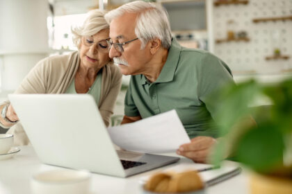 Idosos analisam documento em frente ao laptop, em ambiente doméstico, representando finanças pessoais para aposentados.