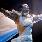 Cantora performa com traje futurista, braços abertos em palco iluminado, transmitindo energia e emoção ao público.