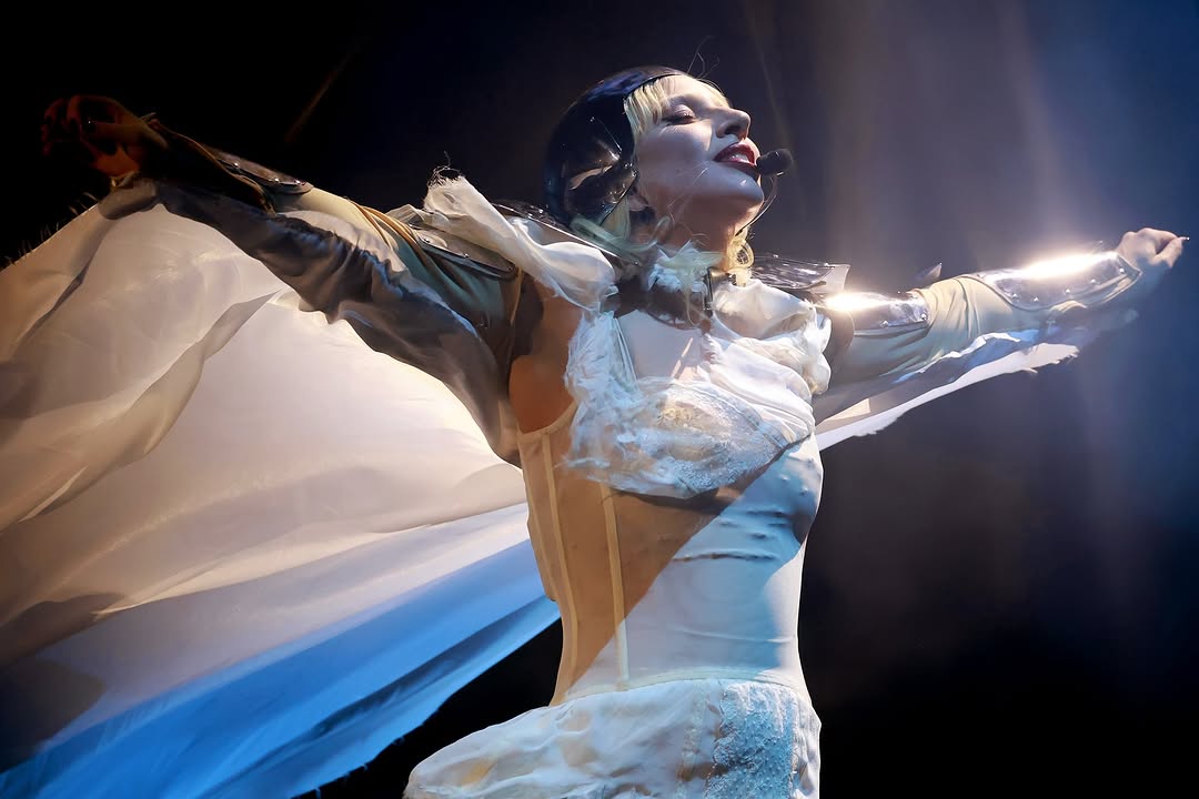 Cantora performa com traje futurista, braços abertos em palco iluminado, transmitindo energia e emoção ao público.