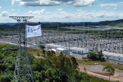 Torres de transmissão elétrica em subestação cercada por vegetação. Pessoa no topo de torre instala bandeira branca.