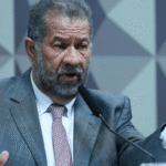 Homem de terno cinza e gravata rosa fala ao microfone gesticulando, em ambiente de paredes geométricas.