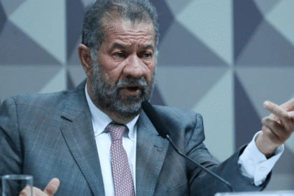 Homem de terno cinza e gravata rosa fala ao microfone gesticulando, em ambiente de paredes geométricas.