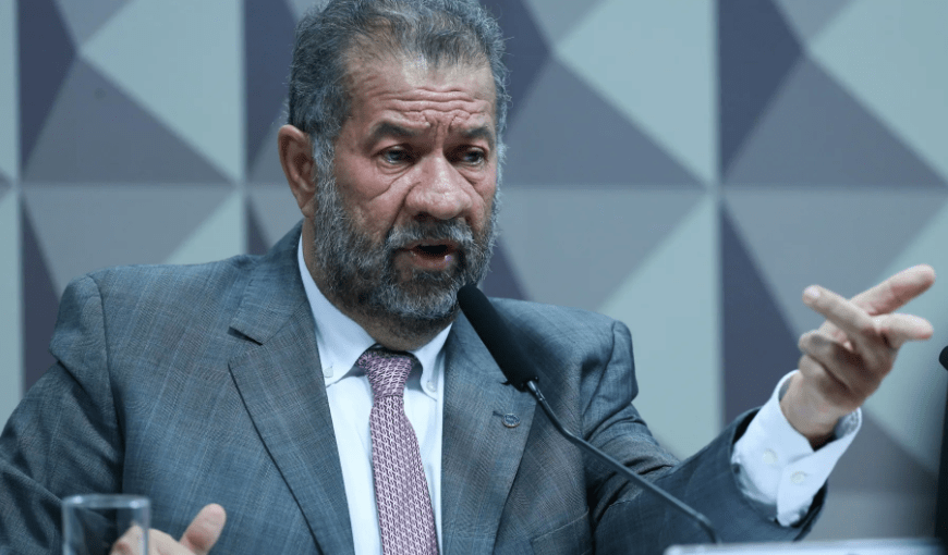 Homem de terno cinza e gravata rosa fala ao microfone gesticulando, em ambiente de paredes geométricas.