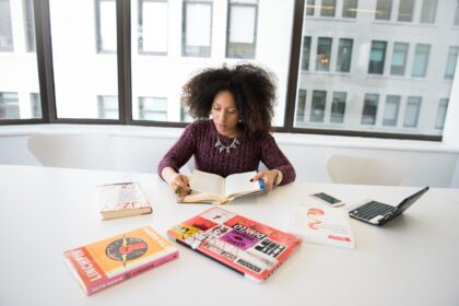 Mulher jovem estudando em um escritório iluminado, cercada por livros e um laptop, anotando em um caderno.