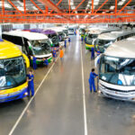 Funcionários em oficina verificam ônibus coloridos enfileirados para manutenção, destacando a indústria automotiva.