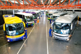 Funcionários em oficina verificam ônibus coloridos enfileirados para manutenção, destacando a indústria automotiva.