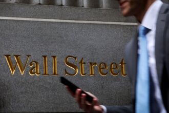 Homem segurando celular passa diante de placa de Wall Street; símbolo financeiro dos Estados Unidos.