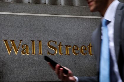 Homem segurando celular passa diante de placa de Wall Street; símbolo financeiro dos Estados Unidos.