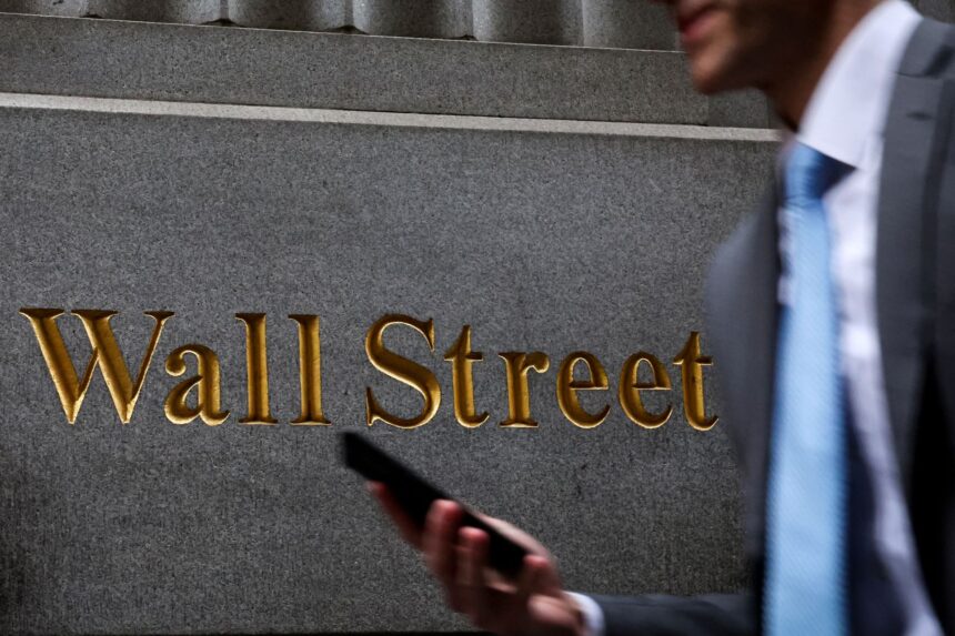 Homem segurando celular passa diante de placa de Wall Street; símbolo financeiro dos Estados Unidos.